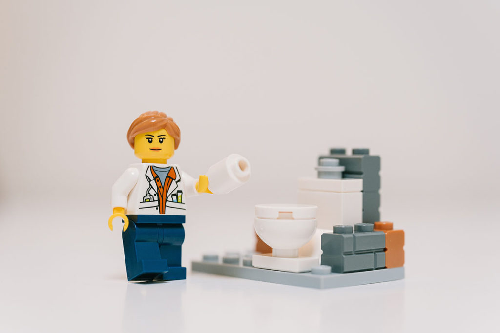 A lego mini figure holding lego toilet paper standing by a lego toilet 